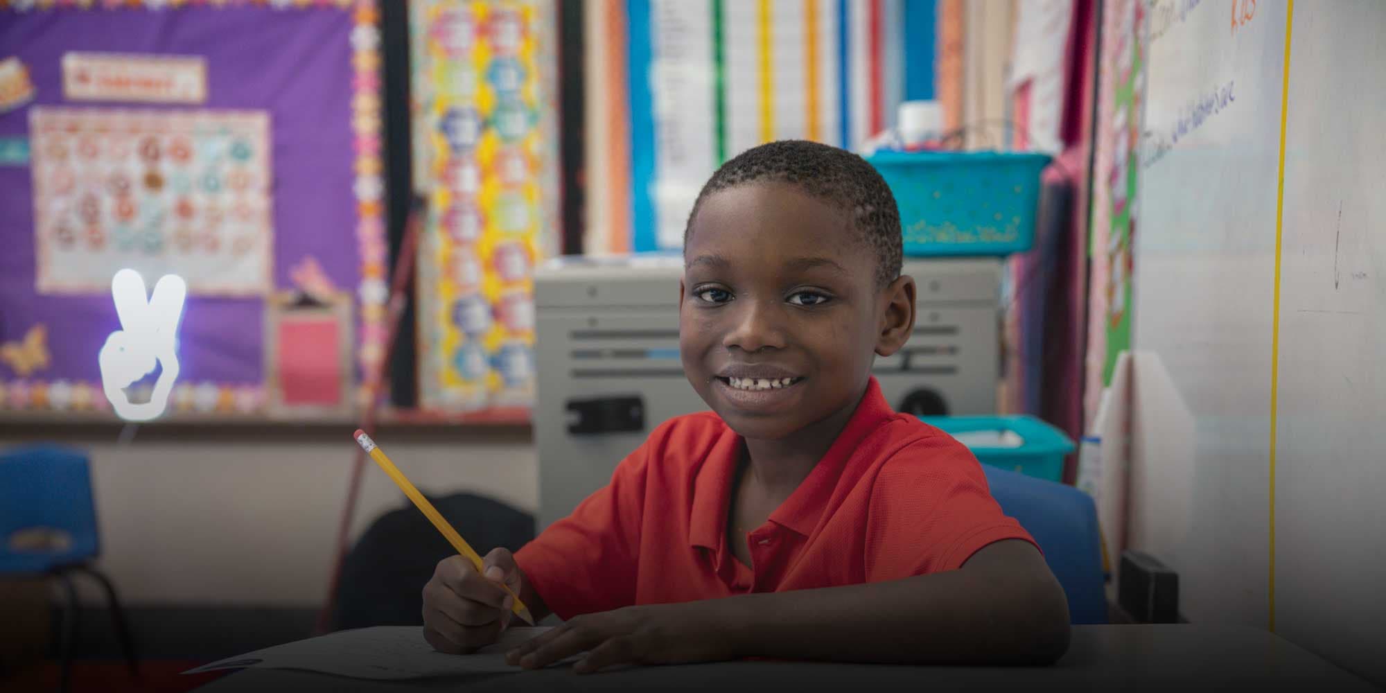 Smiling student in a red shirt writing at a classroom desk.