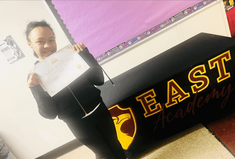 Student smiling, holding a "Talent For Writing" certificate.